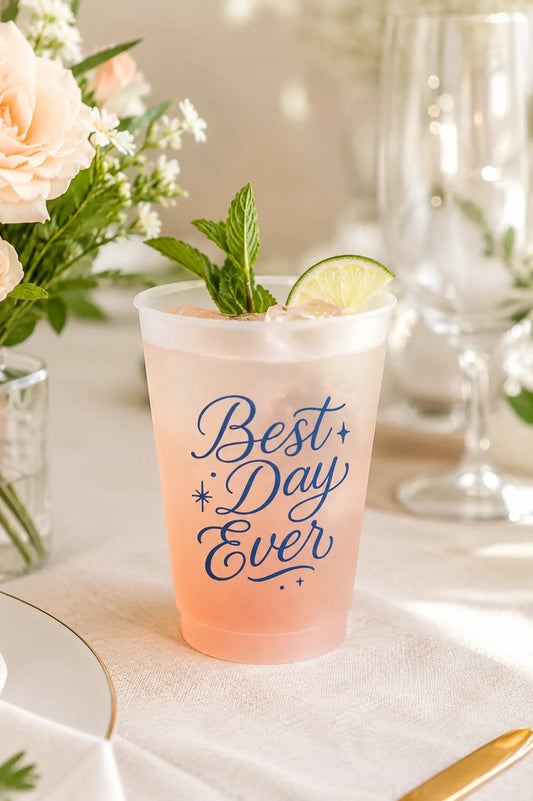 A frosted clear plastic cocktail cup with the phrase 'Best Day Ever' printed in navy blue script, filled with a pale pink drink and garnished with mint and a lime wheel, set on a table with flowers and glassware in the background.