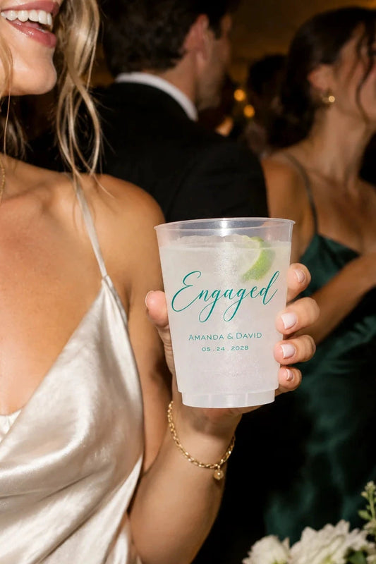 A person in a champagne satin dress holds a clear plastic cocktail cup printed with the word "Engaged" and personalized names and date in teal, at a wedding reception with other guests and candles in the background.