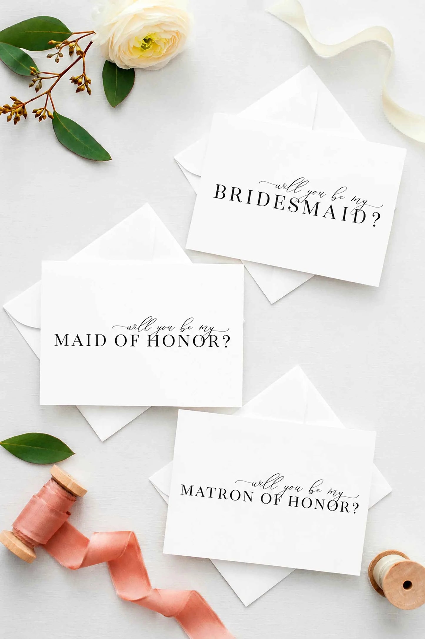 Three white Will you be my proposal cards for a bridal shower or bachelorette party invitation cards with matching envelopes, styled in a flat lay with a white rose, green leaves, pink ribbon, and wooden spools on a light gray background.