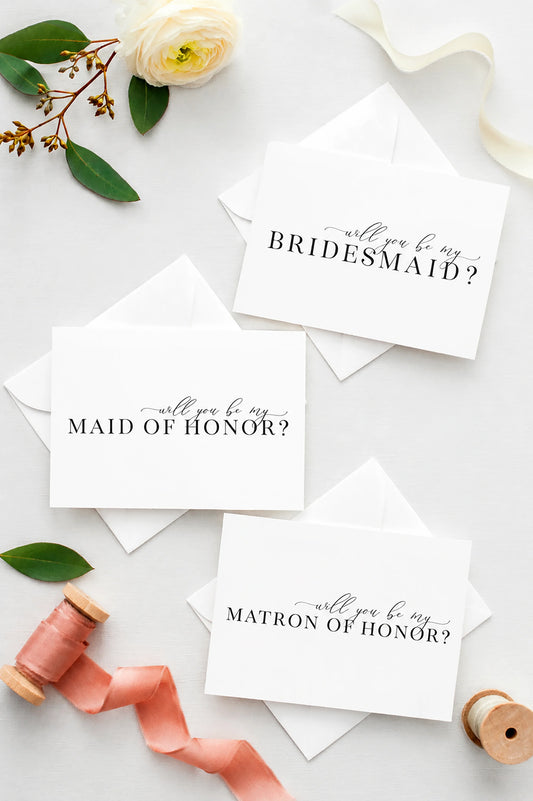 Three white Will you be my proposal cards for a bridal shower or bachelorette party invitation cards with matching envelopes, styled in a flat lay with a white rose, green leaves, pink ribbon, and wooden spools on a light gray background.