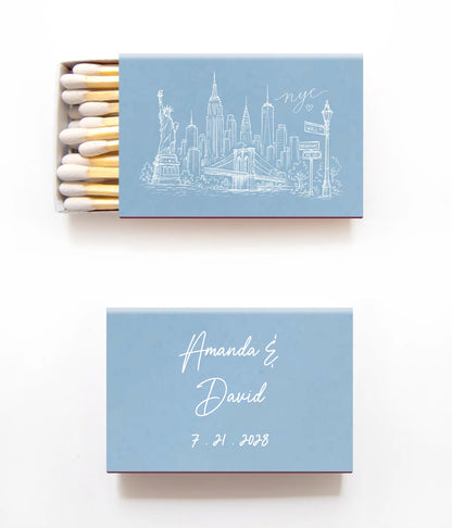 A personalized matchbox set on a clean white background, featuring a light blue cover with a white New York City skyline illustration on the top matchbox and white script names and date on the bottom matchbox.