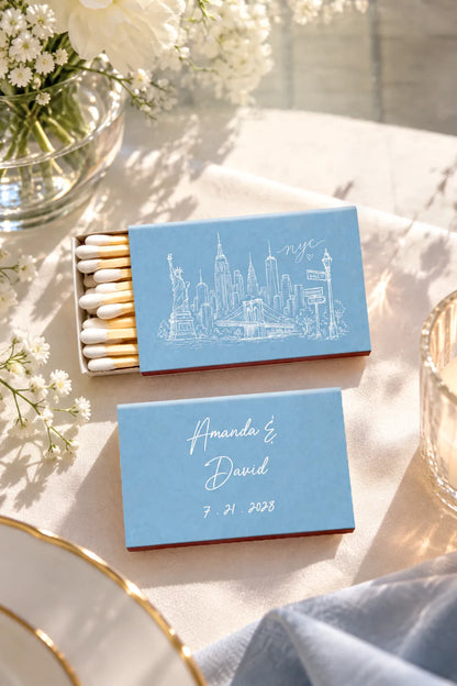 Personalized light blue matchboxes on a sunlit table with white flowers and glassware. One matchbox shows a white New York City skyline illustration with 'nyc' script, and the other has the names 'Amanda & David' and the date '7.