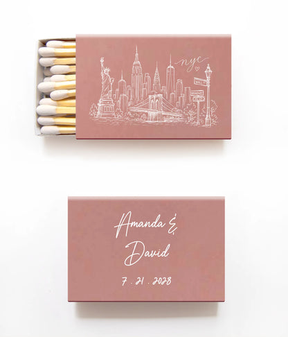 Two dusty rose matchboxes on a white background, one open showing white and gold matches with a white New York City skyline illustration, and one closed with the names 'Amanda & David' and the date '7.21.