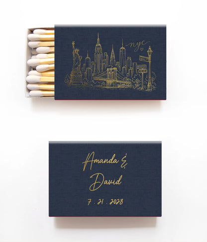 Two navy blue matchboxes with gold foil detailing on a white background. The top matchbox is open, showing white match heads and golden matchsticks, with a gold New York City skyline illustration and the text 'nyc' on the cover.