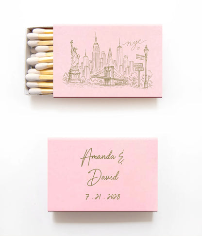 A pink matchbox set on a white background, with one matchbox open showing white and gold matches and one closed printed with a New York City skyline and the text 'nyc' on the lid, plus another closed matchbox below with personalized names 'Amanda & David' and the date '7.21.