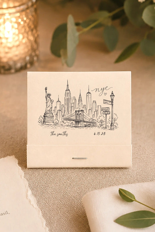 Cream colored matchbooks that have a hand drawn image of the New York city skyline with the names the smiths and their wedding date printed in black ink on a beige wedding table with greenery and a candle 