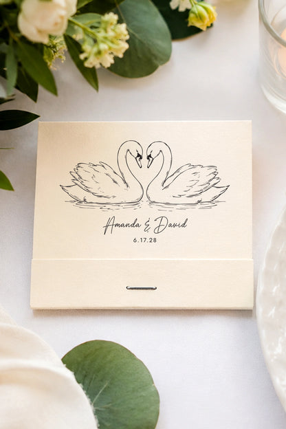 ivory wedding matchbook with swan design and personalized text on a white surface with greenery.