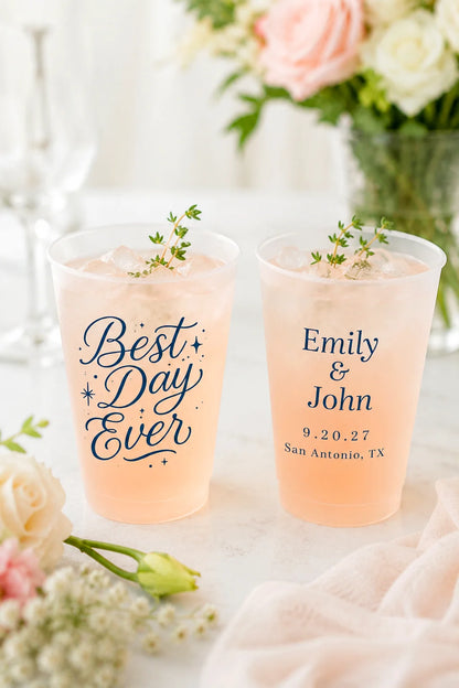 Two frosted plastic cups filled with a pale pink beverage, placed on a light surface with floral decor in the background. One cup reads “Best Day Ever” and the other reads “Emily & John 9.