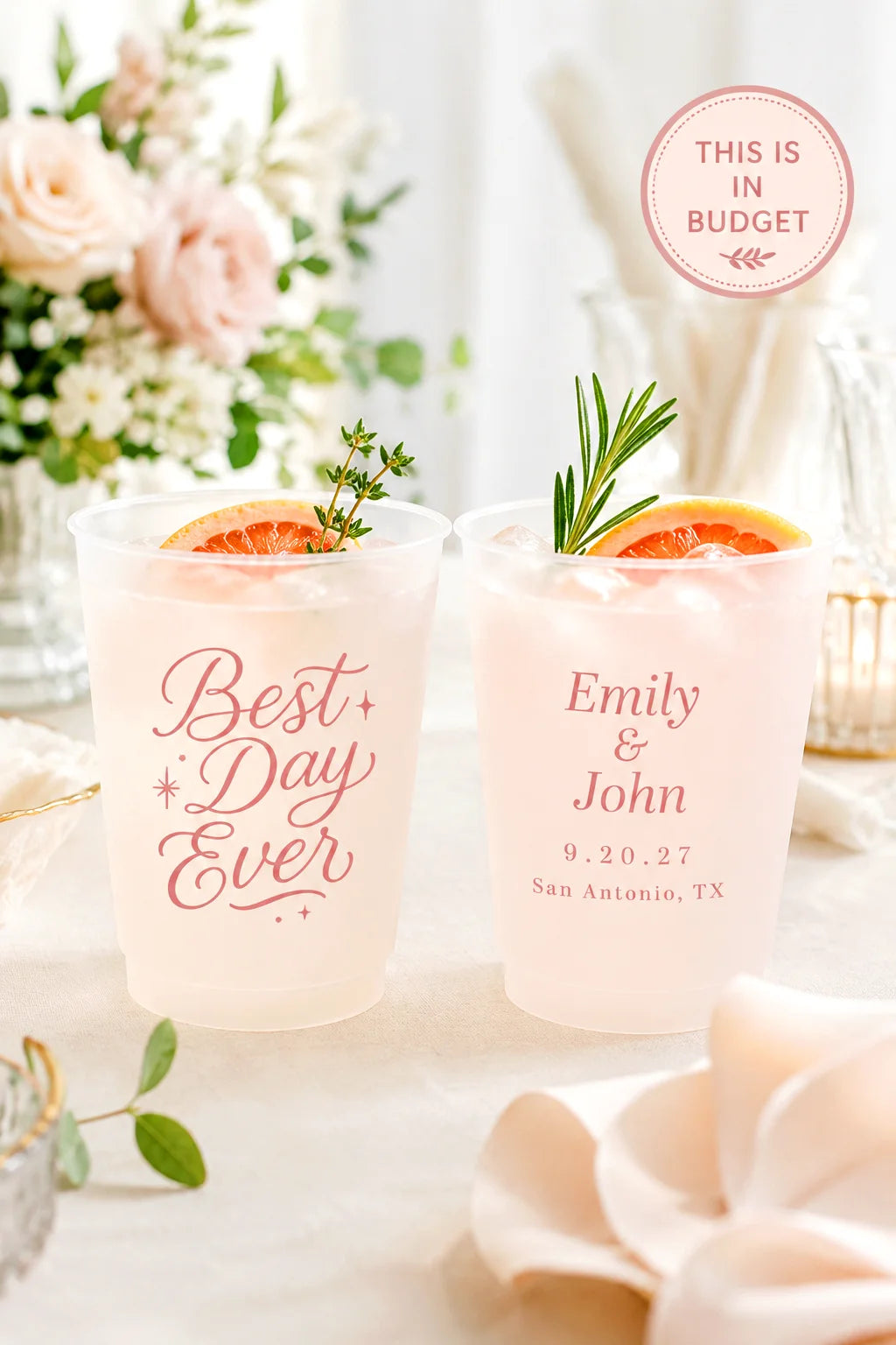 Two clear plastic cocktail cups with pink personalized text, shown filled with a pale pink drink and garnished with orange slices and rosemary on a styled table setting. A pink circular badge reads 'THIS IS IN BUDGET'.