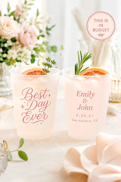 Two clear plastic cocktail cups with pink personalized text, shown filled with a pale pink drink and garnished with orange slices and rosemary on a styled table setting. A pink circular badge reads 'THIS IS IN BUDGET'.