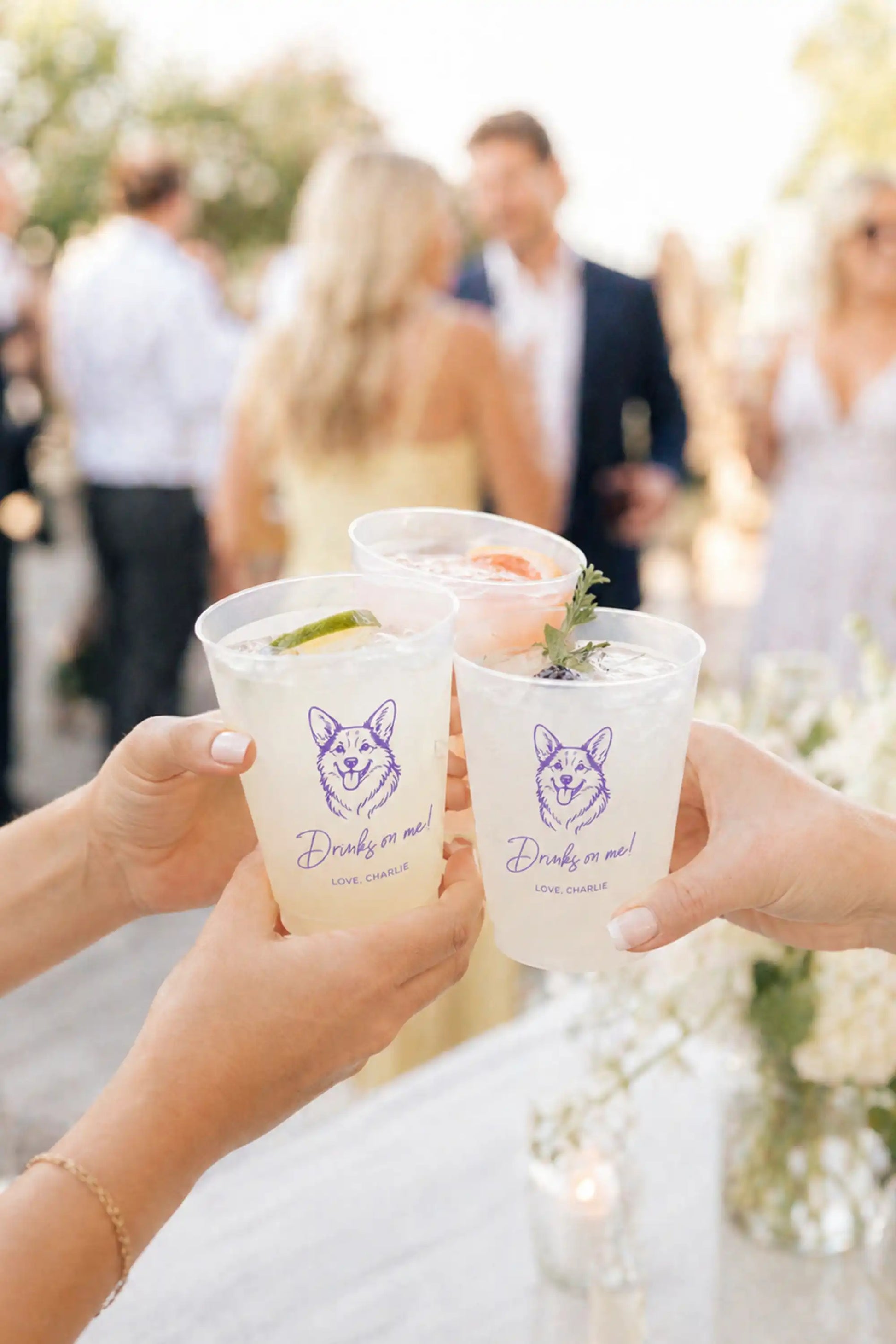 Two hands holding wedding cups with a corgi dog, blurred wedding background