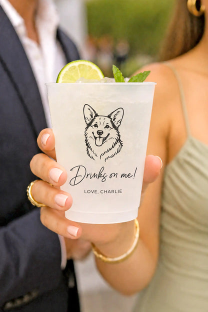 Person holding a frosted cup with a corgi dog illustration and text, garnished with a lime and mint.