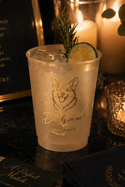 Clear plastic cup with a corgi dog illustration and 'Drinks on me!' text, filled with a drink and garnished with a lime and rosemary sprig, on a dark surface with candles in the background.
