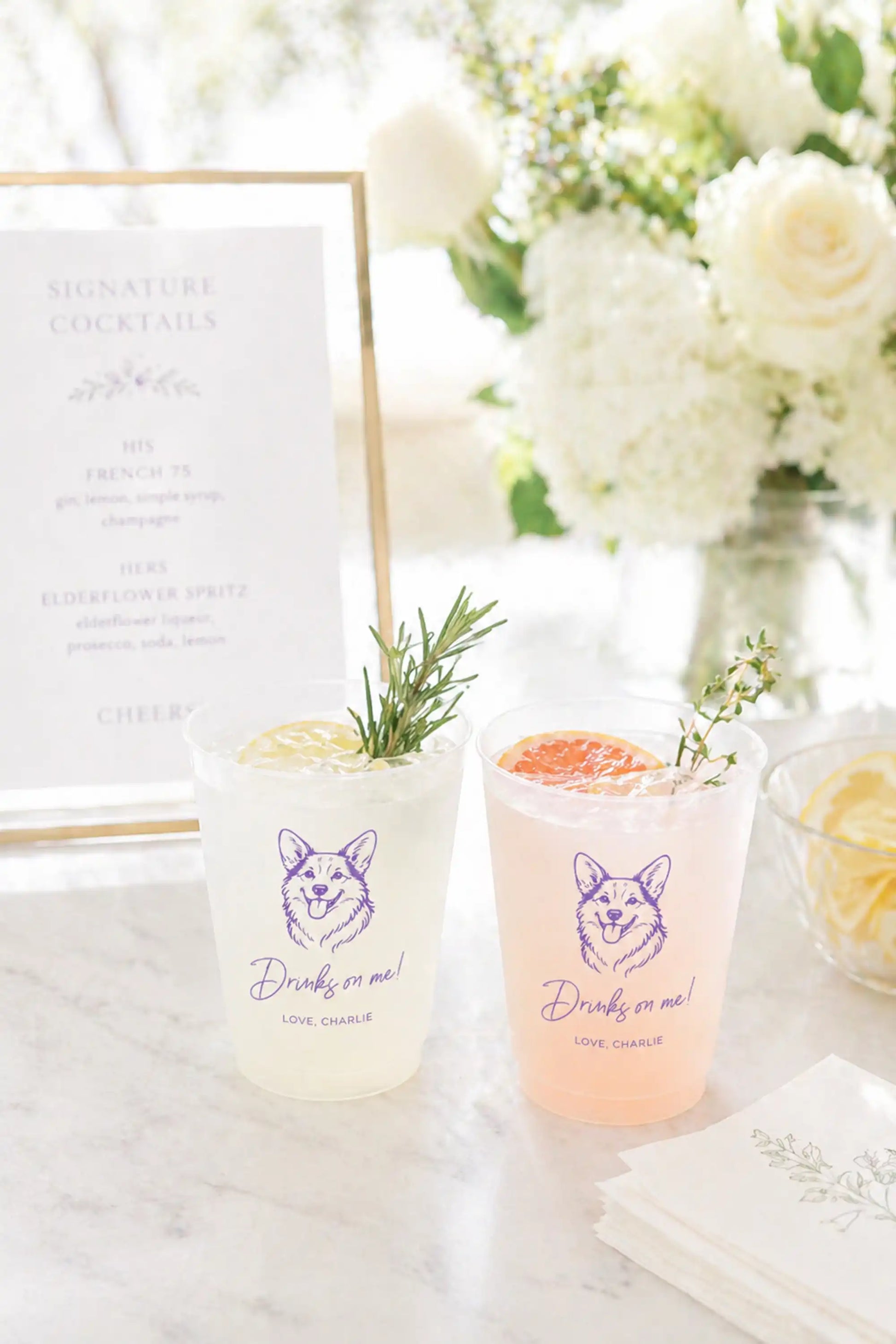 Two cocktails in clear glasses with a corgi dog and Drinks on me text on a marble surface, surrounded by flowers and a cocktail menu.