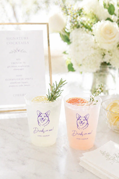 Two cocktails in clear glasses with a corgi dog and Drinks on me text on a marble surface, surrounded by flowers and a cocktail menu.