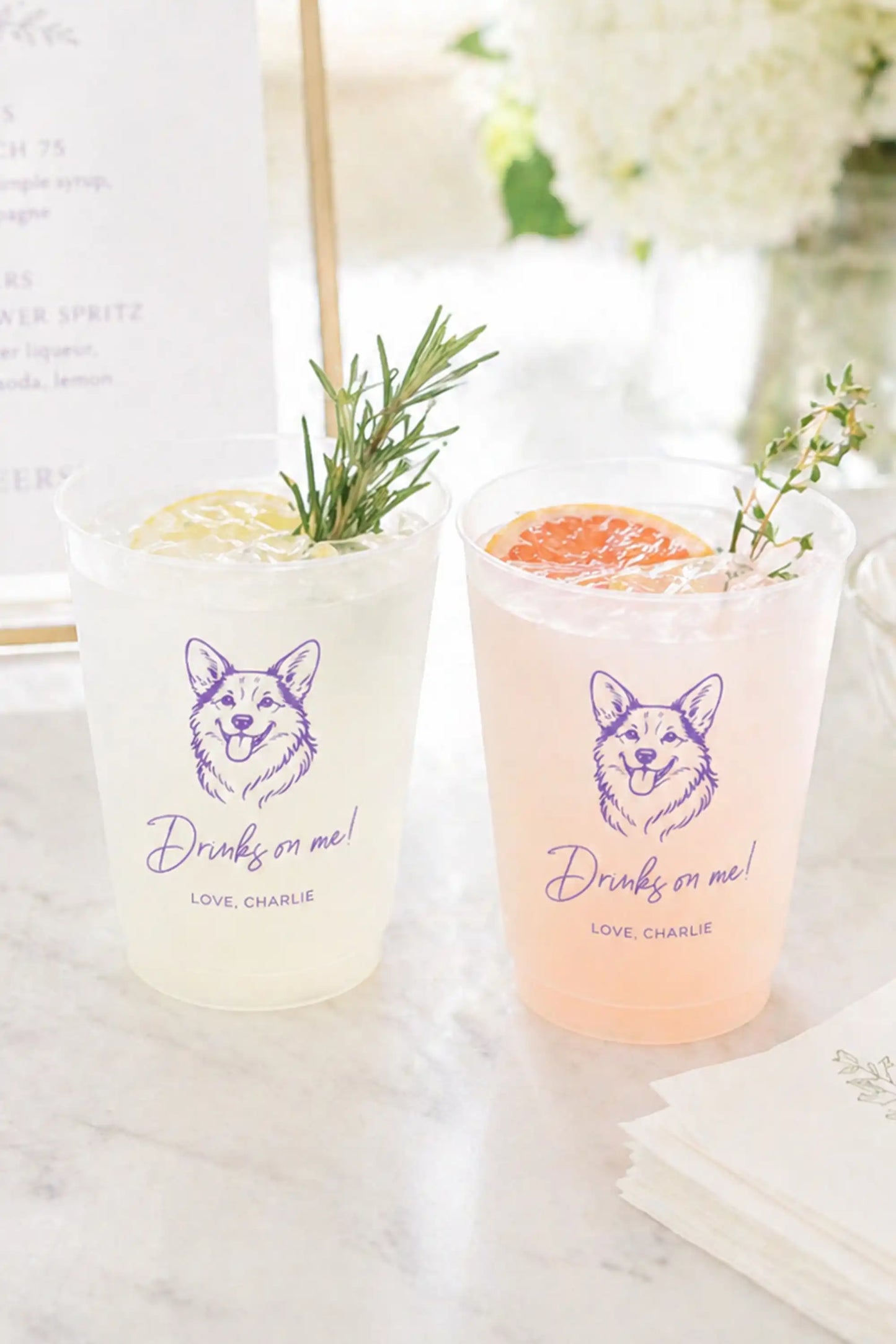 Two cocktails in clear glasses with corgi dog graphic and Drinks on me text on a marble surface.