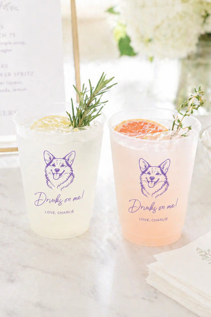 Two cocktails in clear glasses with corgi dog graphic and Drinks on me text on a marble surface.
