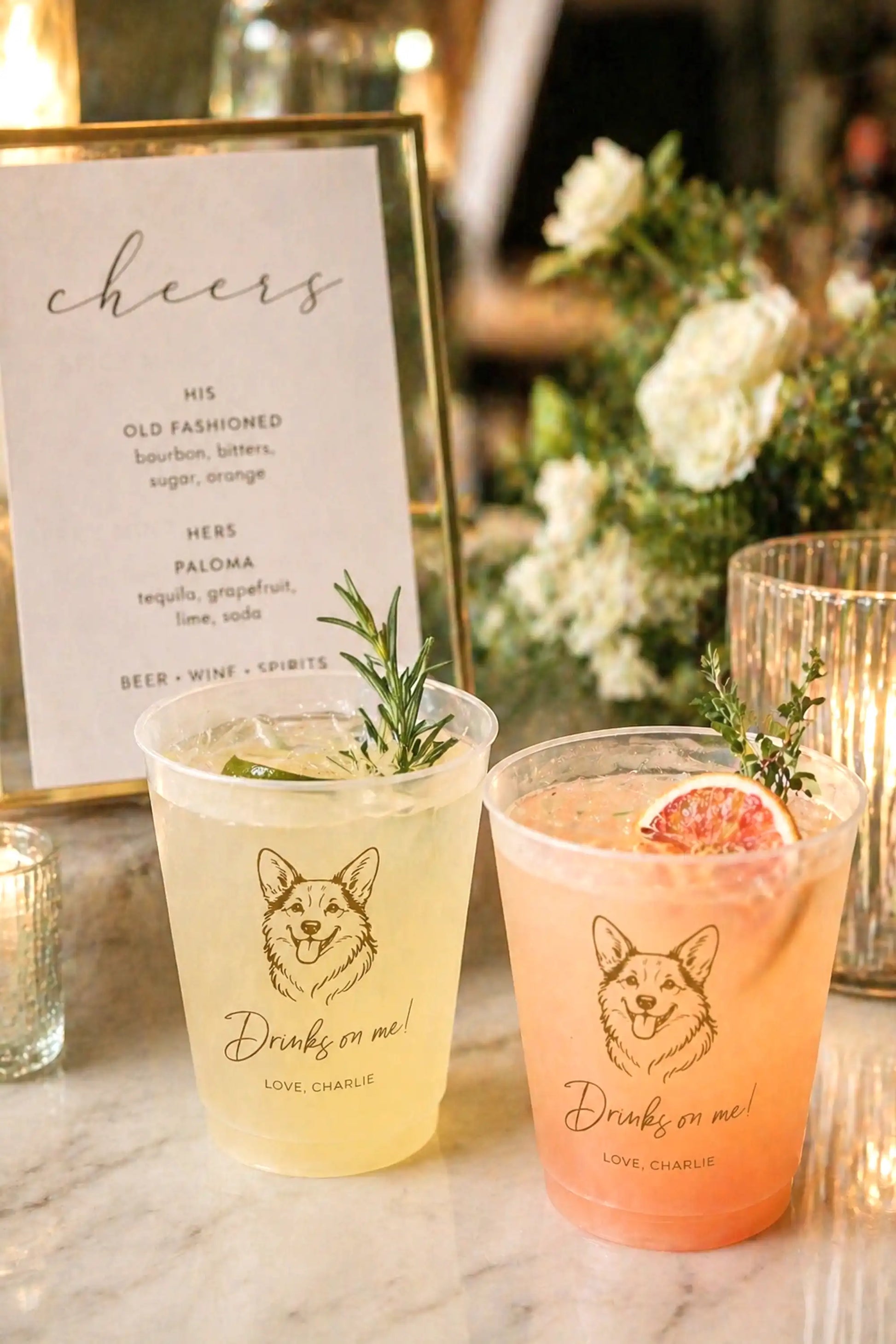Two cocktails in cups with a corgi dog illustrations and Drinks on Me text on a marble surface, accompanied by a 'cheers' sign and floral decorations.