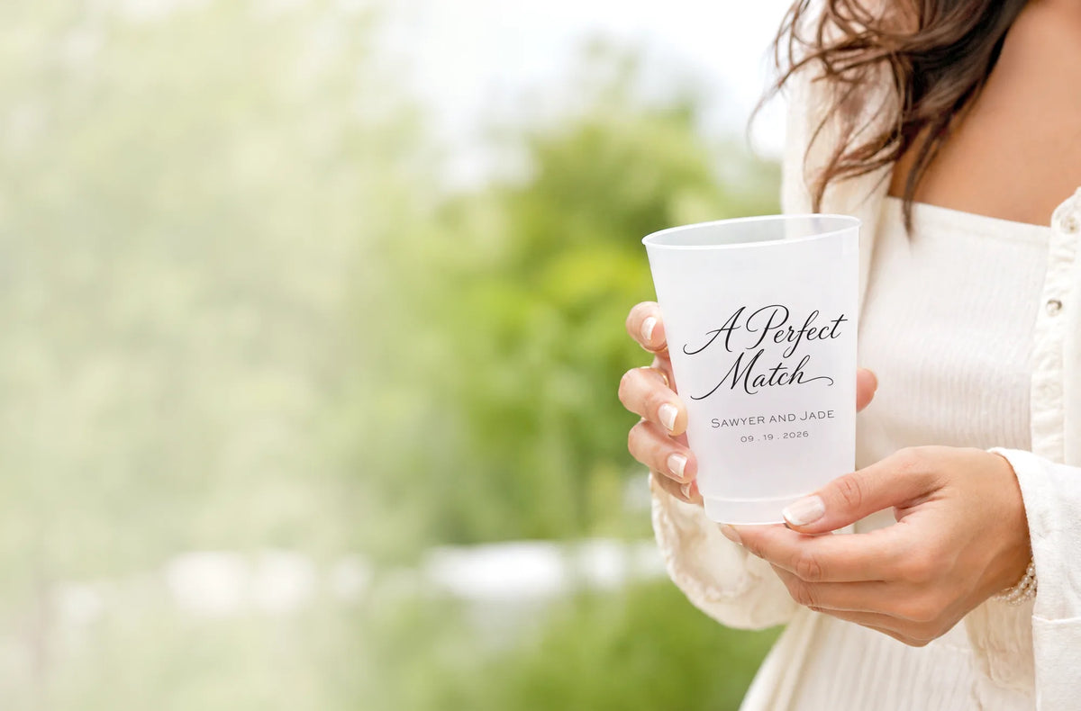 A person in a white off-the-shoulder top holds a frosted white plastic cup with black script text reading 'A Perfect Match' and smaller text 'SANDY AND JADE' on a blurred outdoor background.