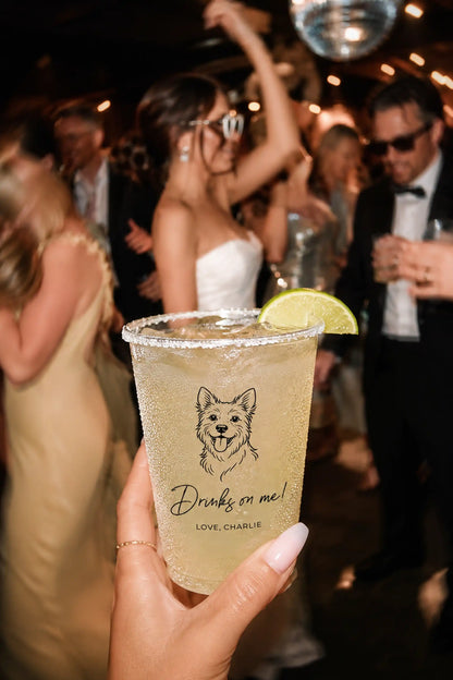 Hand holding a drink with a terrier dog illustration and Drinks on me Love, Charlie text at a wedding social event.
