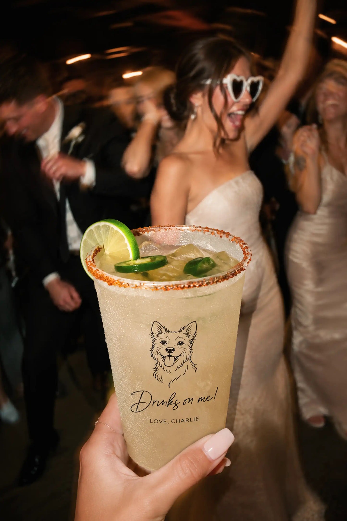 Person holding a margarita with a terrier dog illustration and drinks on me love charlie text, with a bride and groom in the background.