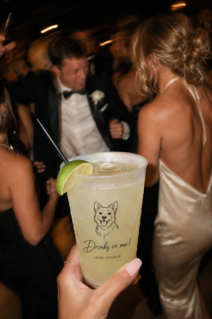 Person holding a drink with a Terrier dog illustration and Drinks on me love charlie text at a wedding social event.
