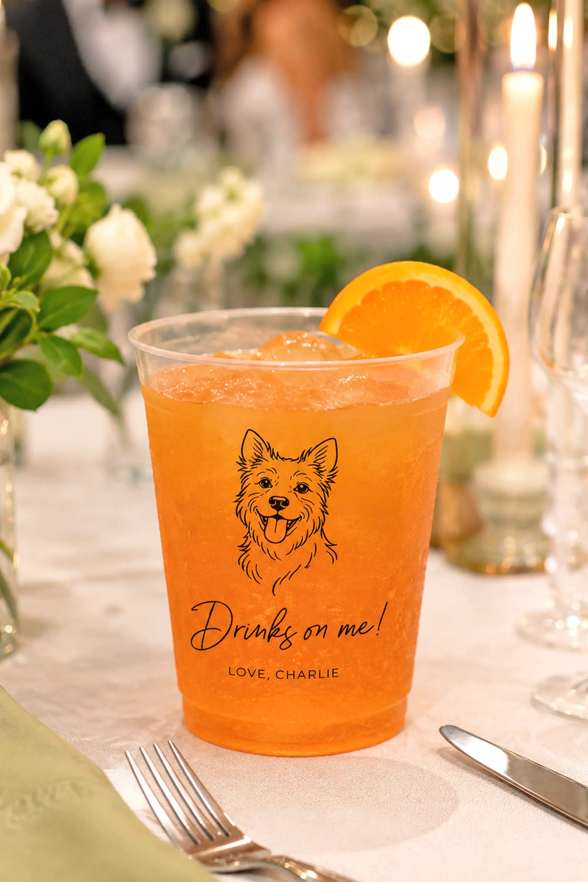 Orange drink in a cup with a terrier dog illustration and drinks on me love charlie wedding text on a table setting.