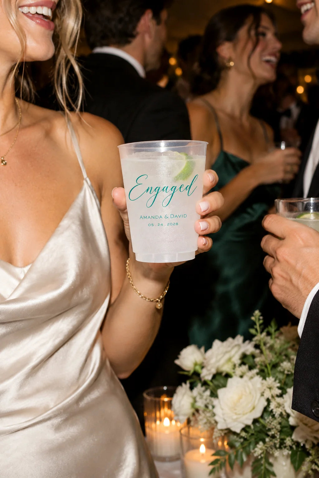 A person in a champagne satin dress holds a clear plastic cocktail cup printed with the word "Engaged" and personalized names and date in teal, at a wedding reception with other guests and candles in the background.