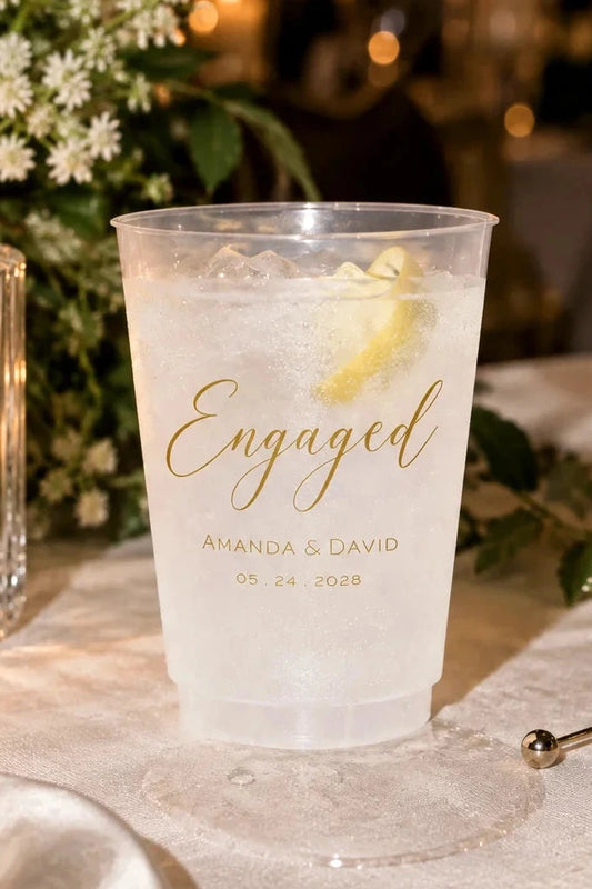 A clear plastic cocktail coupe cup with gold foil text reading 'Engaged Amanda & David 05.24.