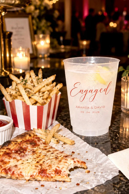 Personalized frosted engagement party cup with custom names and date beside pizza and fries at a late-night wedding snack bar