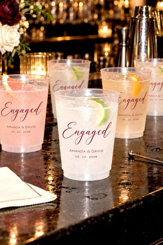 Personalized frosted engagement party cups with custom names and date displayed with cocktails at a candlelit bar