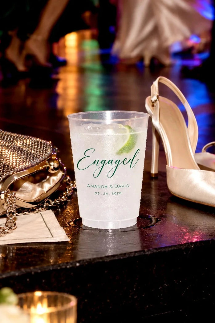Personalized frosted engagement party cup with custom names and date beside heels and a clutch near the dance floor
