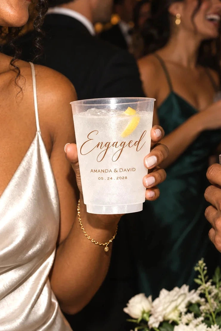 A person in a champagne satin dress holds a clear plastic cocktail cup printed with the word "Engaged" and personalized names and date in teal, at a wedding reception with other guests and candles in the background.
