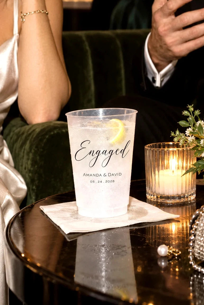 Personalized frosted engagement party cup with custom names and date beside a candle at an elegant cocktail reception