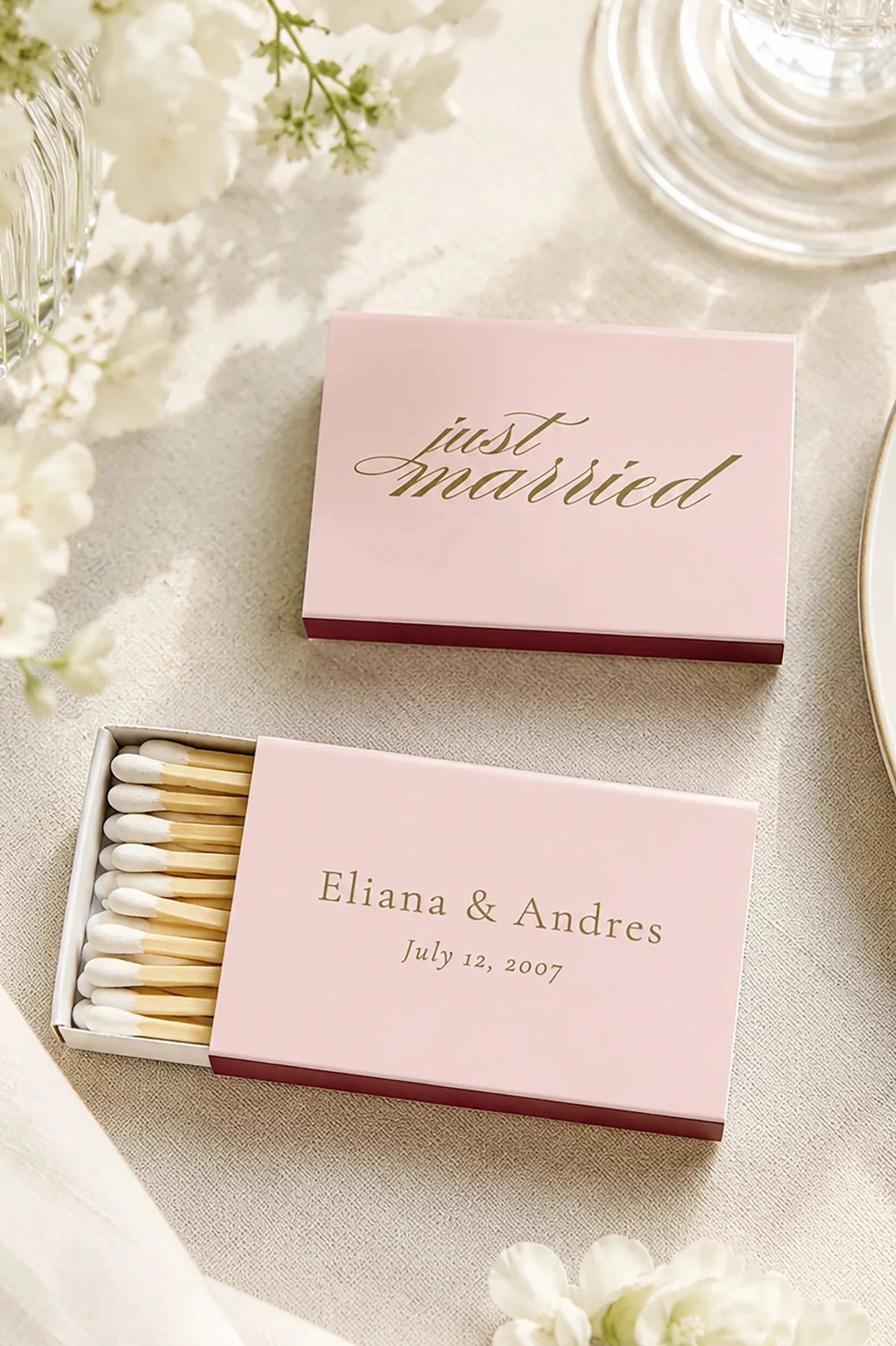 blush pink matchboxes on a ivory linen floral wedding table with a plate and glass. the matches say just married in gold and also have the bride and grooms names and wedding date on the back.