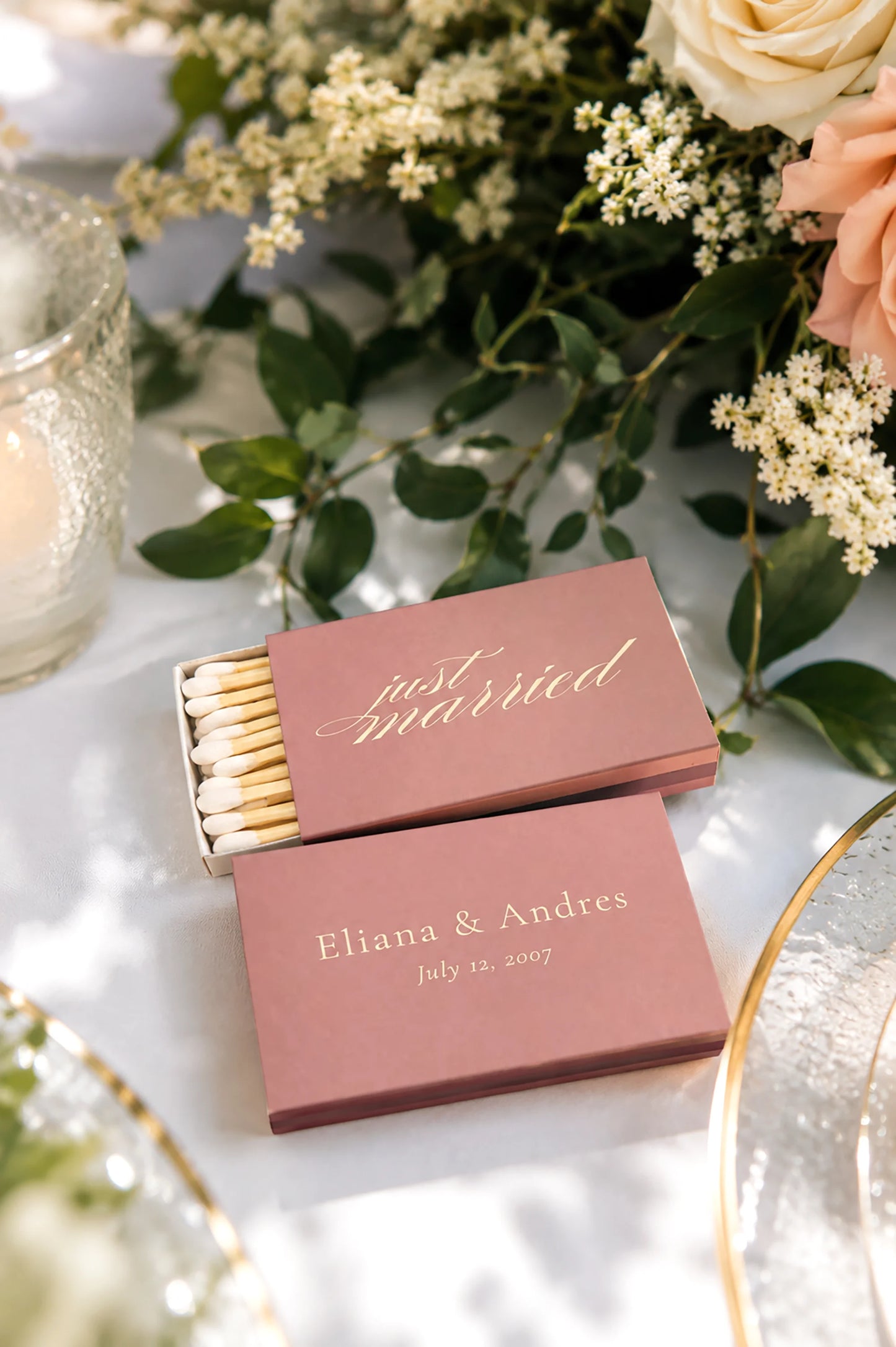 dusy rose matchboxes on a floral wedding table with a plate and candle the matches  say just married in gold and also have the bride and grooms names and wedding date on the back in ivory text.
