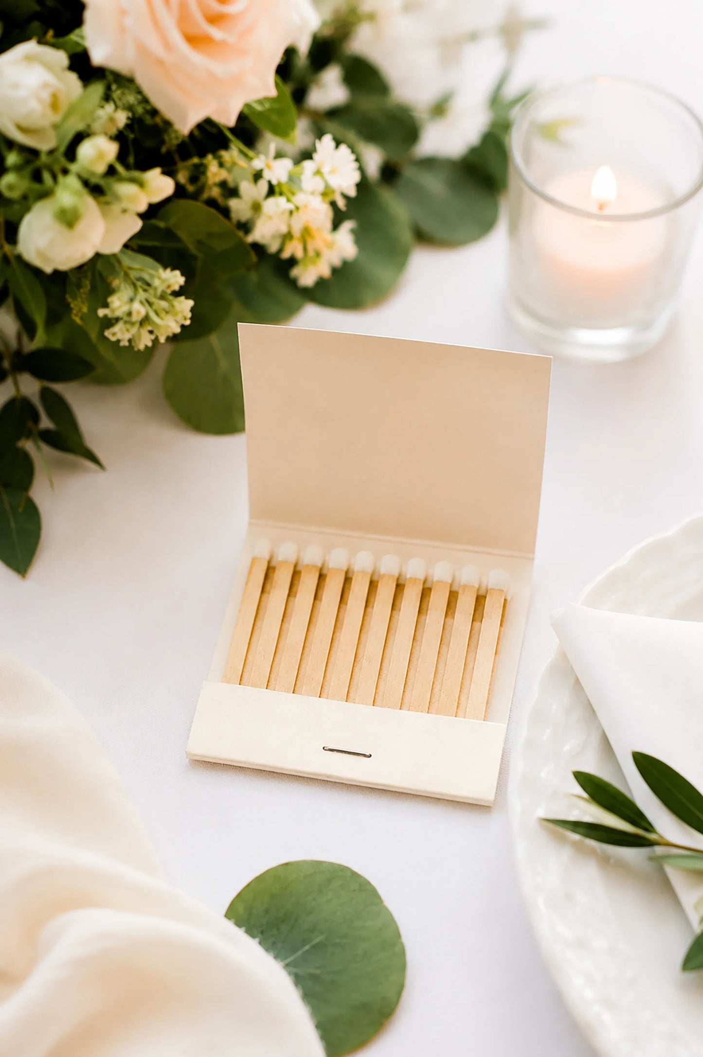 open ivory matchbook and matchsticks on a white wedding table near greenery and roses and a candle. 