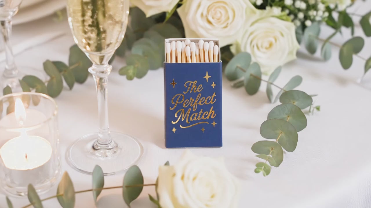 Load video: Close-up video of custom navy blue wedding matchboxes with shimmering gold foil 'The Perfect Match' script. Personalized wedding favors and elegant guest gifts for a 2026 celebration.