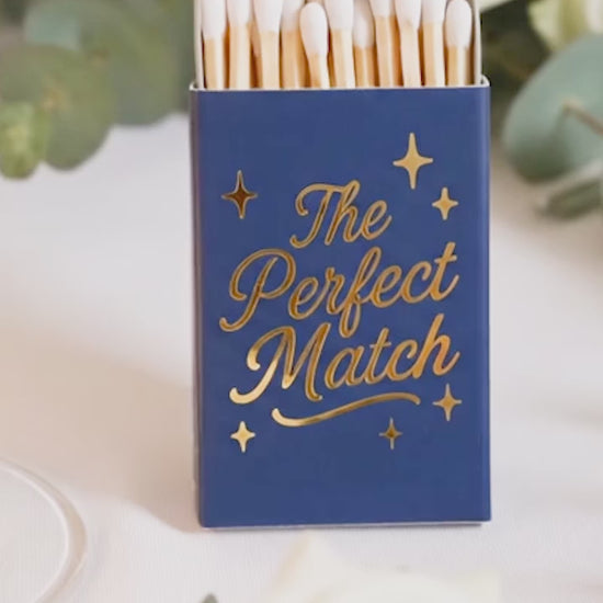 Close-up video of custom navy blue wedding matchboxes with shimmering gold foil 'The Perfect Match' script. Personalized wedding favors and elegant guest gifts for a 2026 celebration.