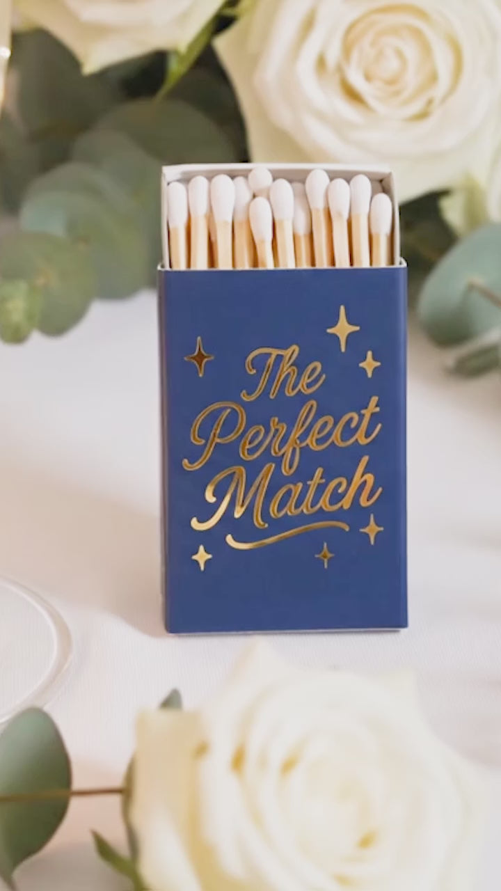 Close-up video of custom navy blue wedding matchboxes with shimmering gold foil 'The Perfect Match' script. Personalized wedding favors and elegant guest gifts for a 2026 celebration.