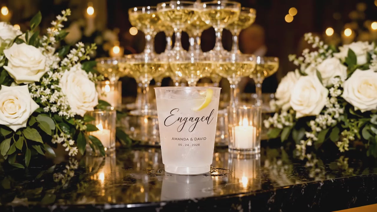 Video of a personalized frosted engagement party cup with custom names and date displayed at a candlelit champagne bar