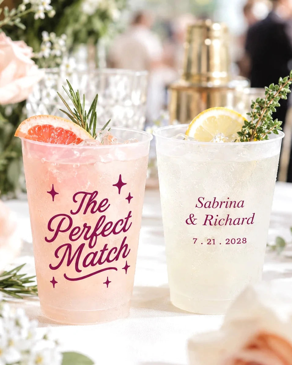 Two customized clear plastic cocktail cups on a white tablecloth, decorated with rosemary and citrus slices. One cup is filled with pink drink and printed with 'The Perfect Match' in burgundy script, while the other is filled with a pale yellow drink and printed with 'Sabrina & Richard 7.