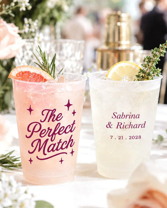 Two customized clear plastic cocktail cups on a white tablecloth, decorated with rosemary and citrus slices. One cup is filled with pink drink and printed with 'The Perfect Match' in burgundy script, while the other is filled with a pale yellow drink and printed with 'Sabrina & Richard 7.