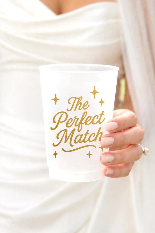 A frosted white plastic party cup with gold foil text reading 'The Perfect Match' and small gold stars, held by a person wearing a white dress.